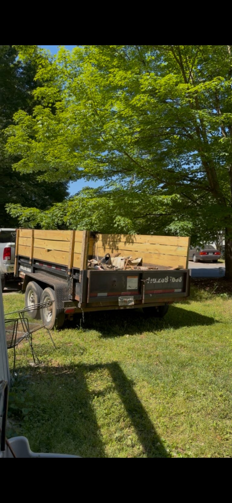 Wooden utility trailer parked on grass beneath green leafy trees on a sunny day