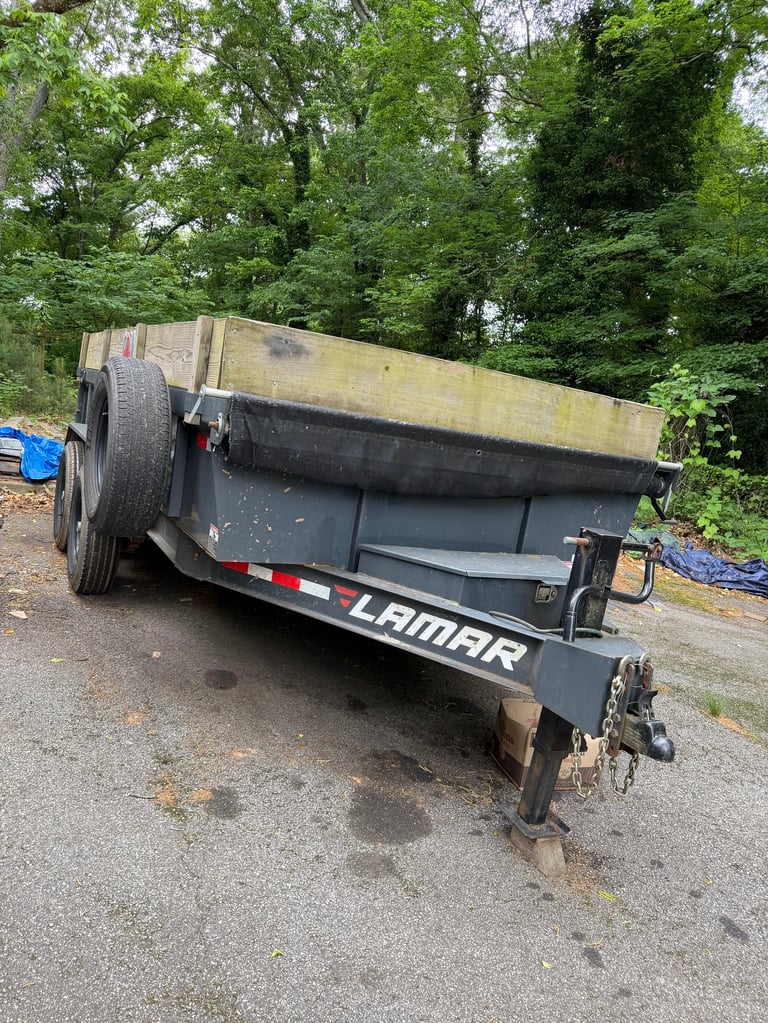 Black Lamar utility trailer with yellow weathered bed parked on asphalt against green trees background