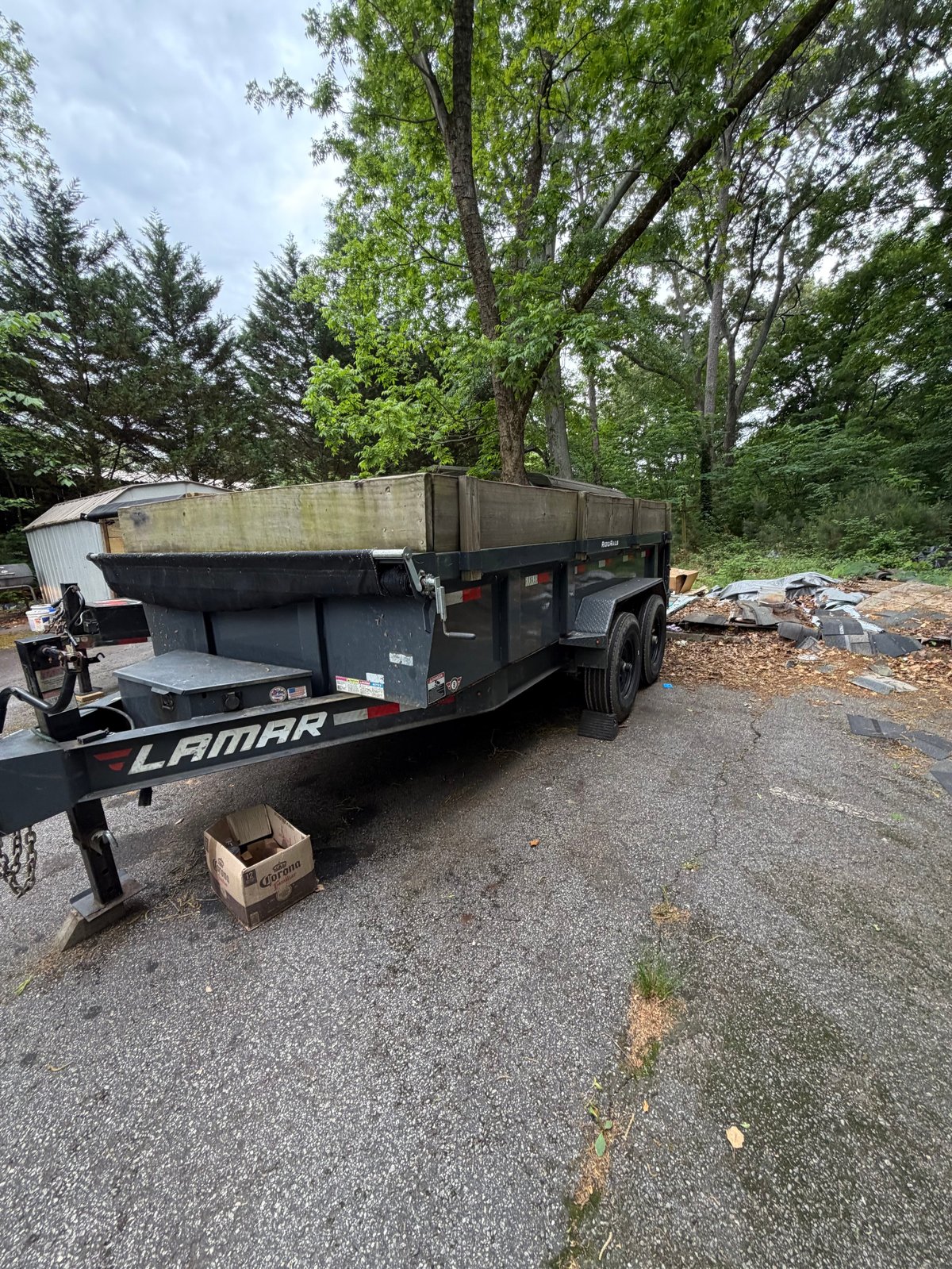 Black Lamar dump trailer parked on asphalt with tall trees in background on a sunny day